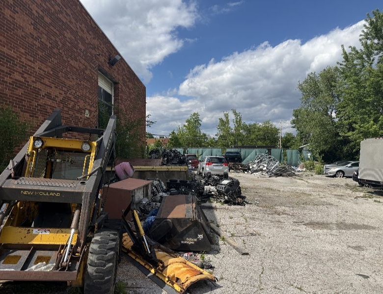 Industrial scrapyard scene with a yellow New Holland forklift near a brick building, and piles of scrap metal and vehicles under a blue sky.