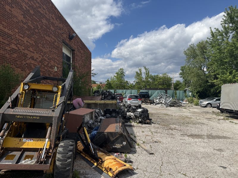 Industrial scrapyard scene with a yellow New Holland forklift near a brick building, and piles of scrap metal and vehicles under a blue sky.