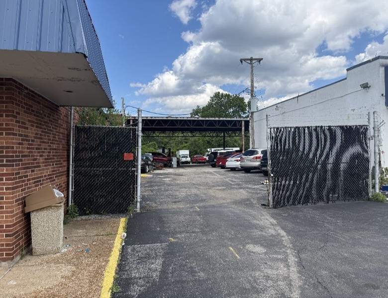 Open chain-link gates lead to a sunlit parking lot with several parked cars under a metal carport, flanked by brick and white buildings.