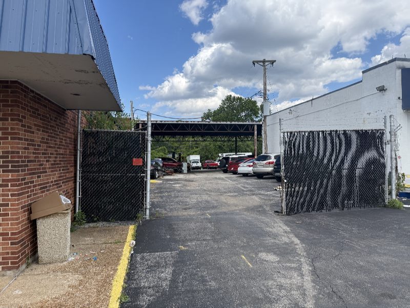Open chain-link gates lead to a sunlit parking lot with several parked cars under a metal carport, flanked by brick and white buildings.