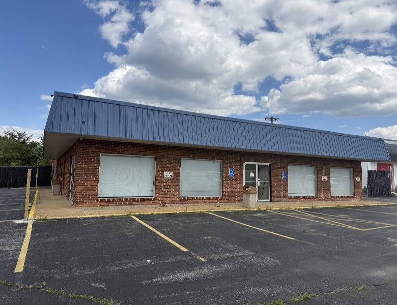 One-story brick commercial building with a blue metal roof, boarded storefront windows, and an empty parking lot in front under a cloudy sky.