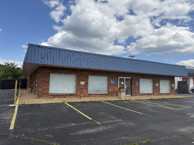 One-story brick commercial building with a blue metal roof, boarded storefront windows, and an empty parking lot in front under a cloudy sky.