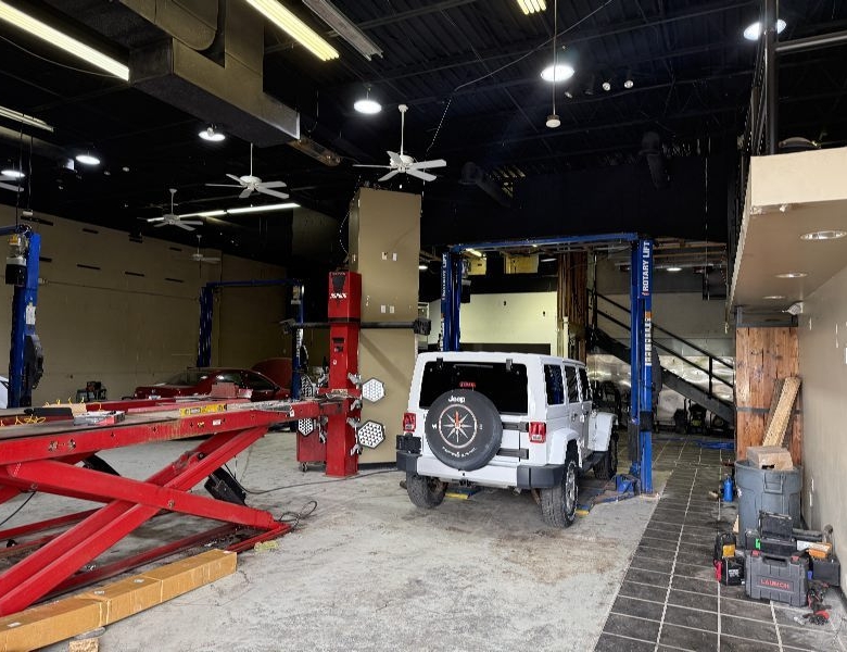 Interior of an auto repair shop with a white Jeep SUV on a hydraulic lift, red car jack arms on the left, and tools along the right wall