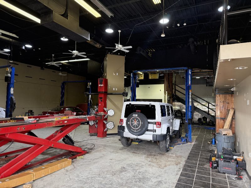 Interior of an auto repair shop with a white Jeep SUV on a hydraulic lift, red car jack arms on the left, and tools along the right wall