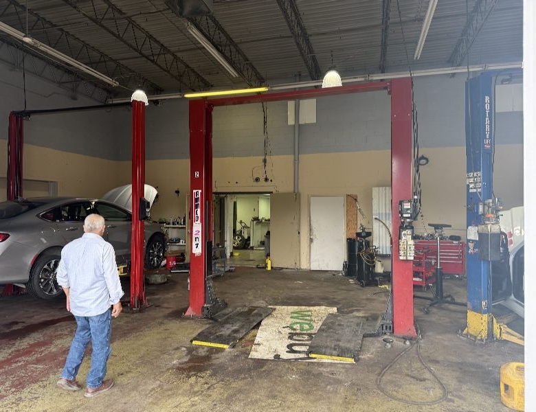 Older man in a light-blue shirt and jeans walks toward a gray car raised on a two-post lift inside an auto repair shop.