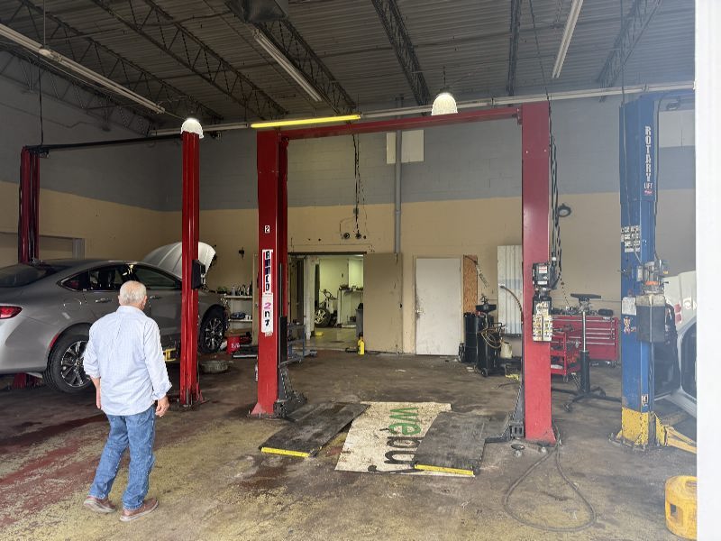 Older man in a light-blue shirt and jeans walks toward a gray car raised on a two-post lift inside an auto repair shop.