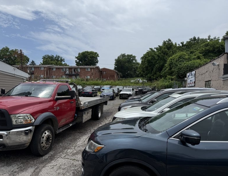 Red tow truck with a flatbed beside a line of sedans in a gravel car lot, brick apartments in the background under a partly cloudy sky.