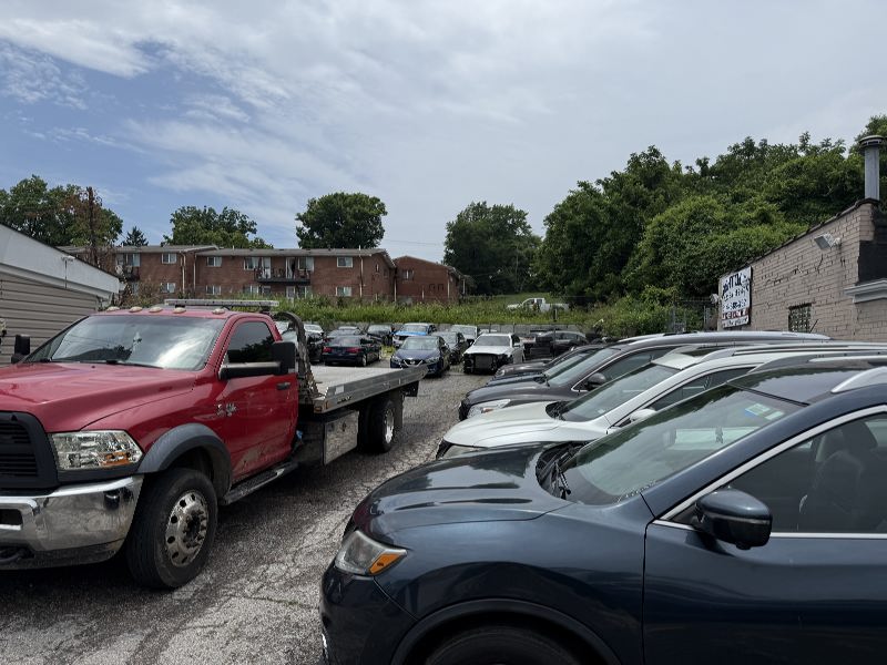 IMG_7794 Red tow truck with a flatbed beside a line of sedans in a gravel car lot, brick apartments in the background under a partly cloudy sky.