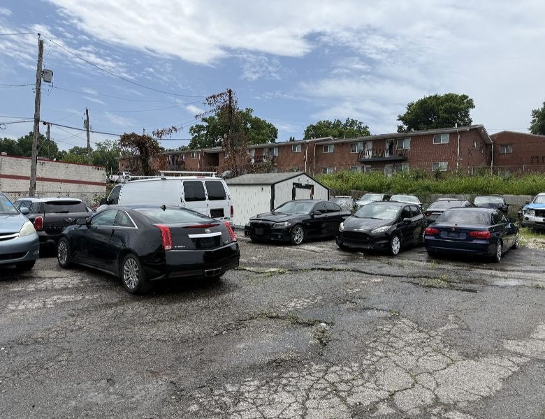 Row of parked cars in a cracked asphalt lot with a white storage shed and brick apartment buildings in the background under a partly cloudy sky.