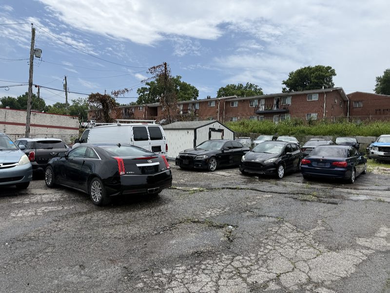 IMG_7795 Row of parked cars in a cracked asphalt lot with a white storage shed and brick apartment buildings in the background under a partly cloudy sky.