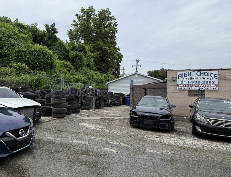 Used car lot with several cars in front of a small building and piles of tires along a fence.