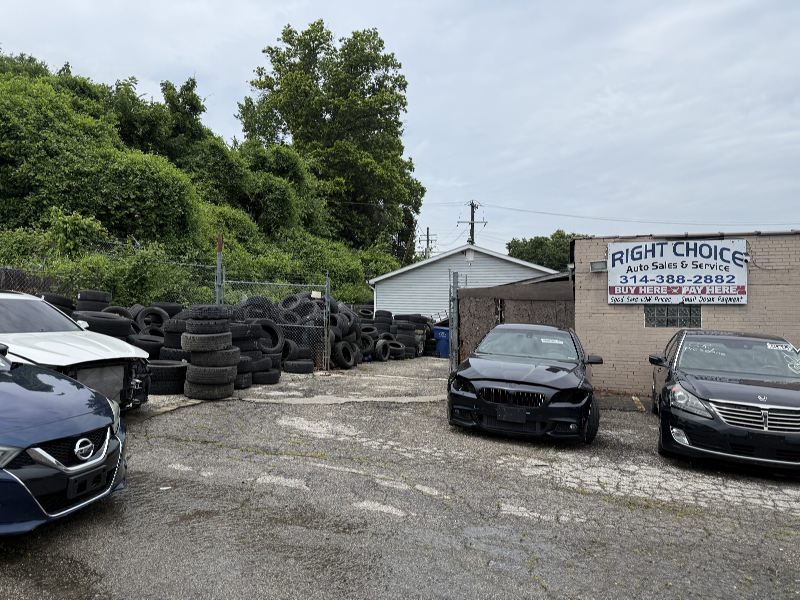 IMG_7796 Used car lot with several cars in front of a small building and piles of tires along a fence.