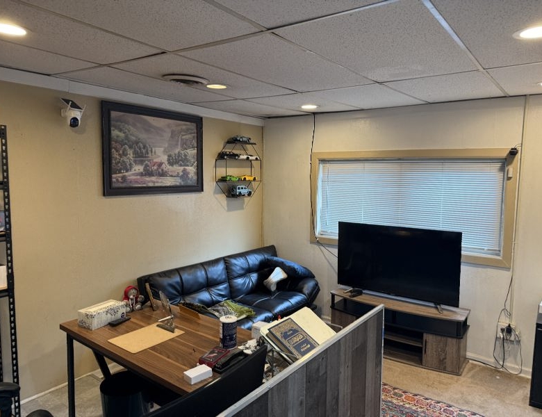Cozy basement living space with a black leather sofa, wooden desk cluttered with items, and a flat-screen TV on a stand by a windowed wall.