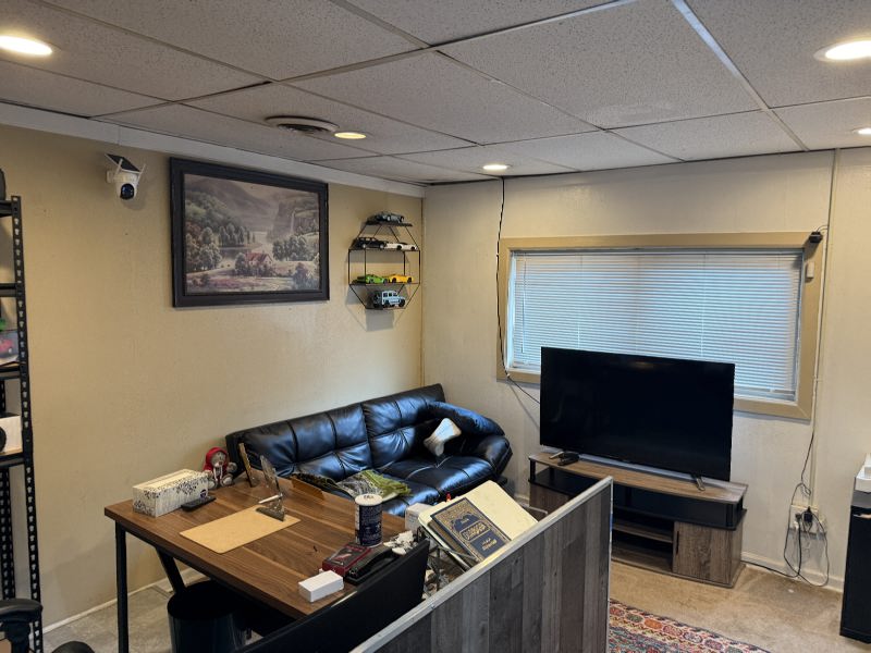 IMG_7798 Cozy basement living space with a black leather sofa, wooden desk cluttered with items, and a flat-screen TV on a stand by a windowed wall.