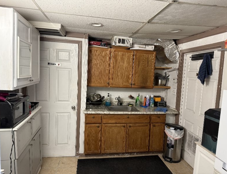 Basement kitchenette with wooden cabinets, a sink, dish rack, and cleaning supplies on a granite counter; a door labeled Private on the left and a trash can on the right.