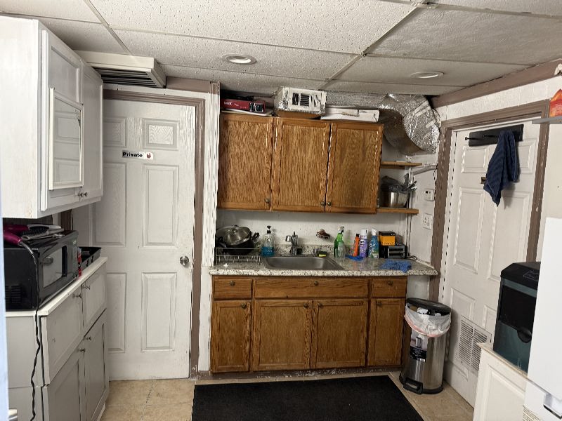 IMG_7799 Basement kitchenette with wooden cabinets, a sink, dish rack, and cleaning supplies on a granite counter; a door labeled Private on the left and a trash can on the right.