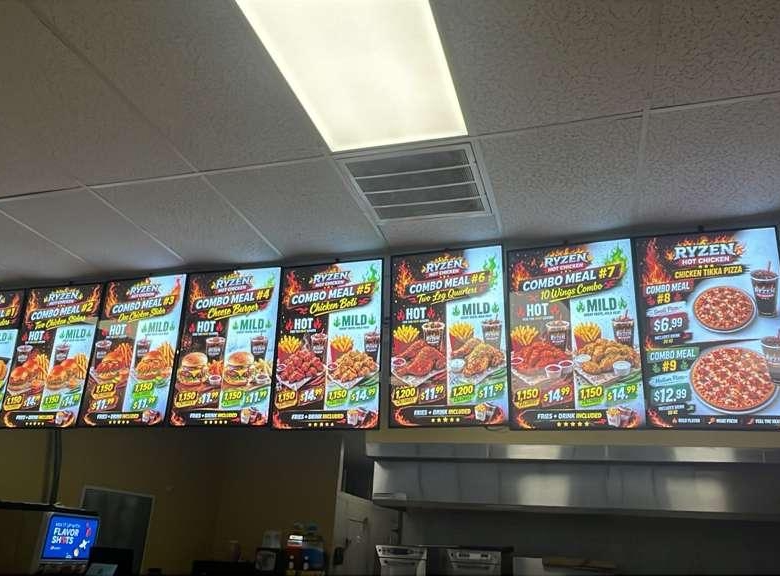 Row of illuminated digital menu boards above a fast-food counter showing combo meals, chicken, pizza, and sides with prices on each board.
